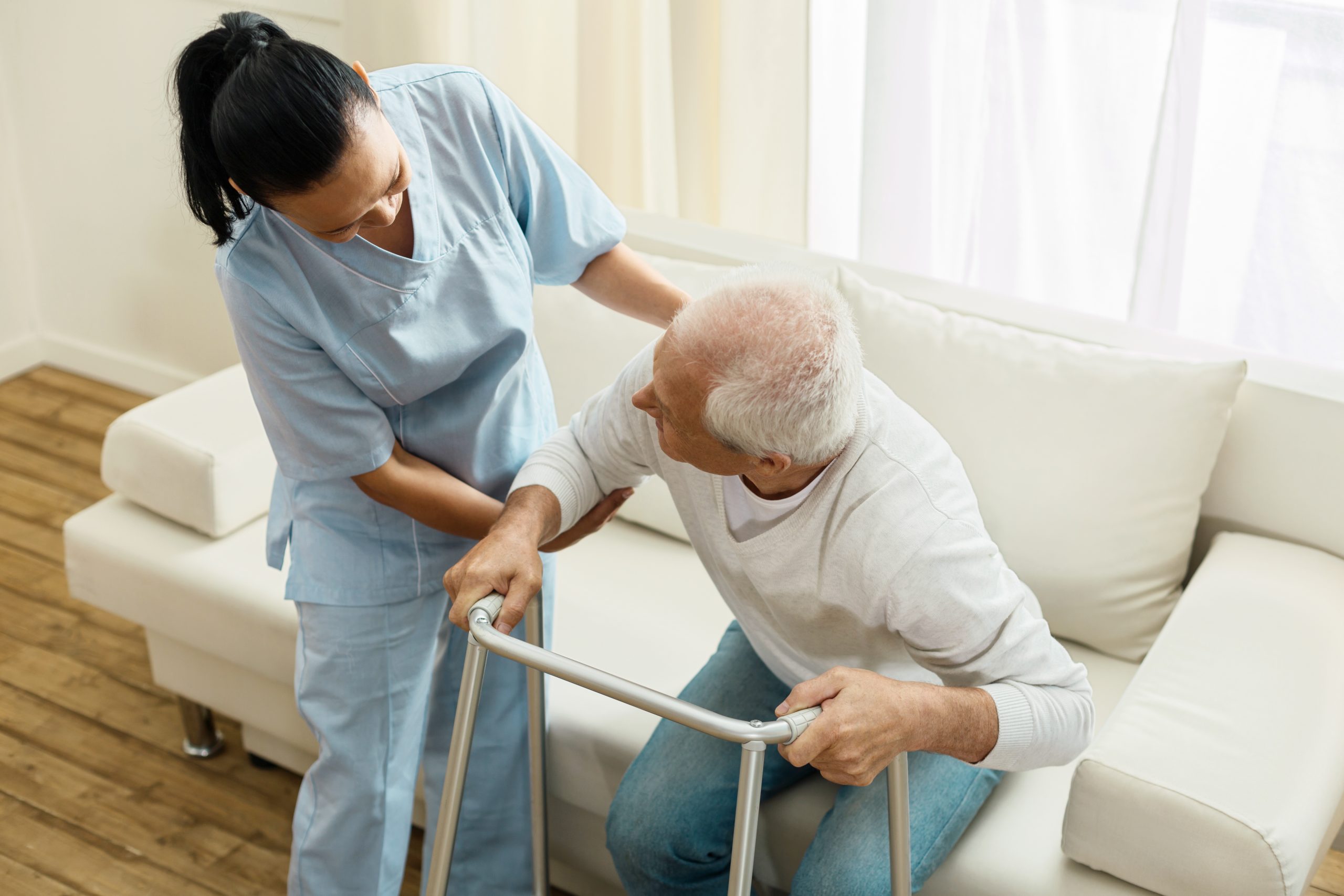 Caring about people. Pleasant delighted friendly caregiver holding her elderly patient and helping him to stand up while doing her job