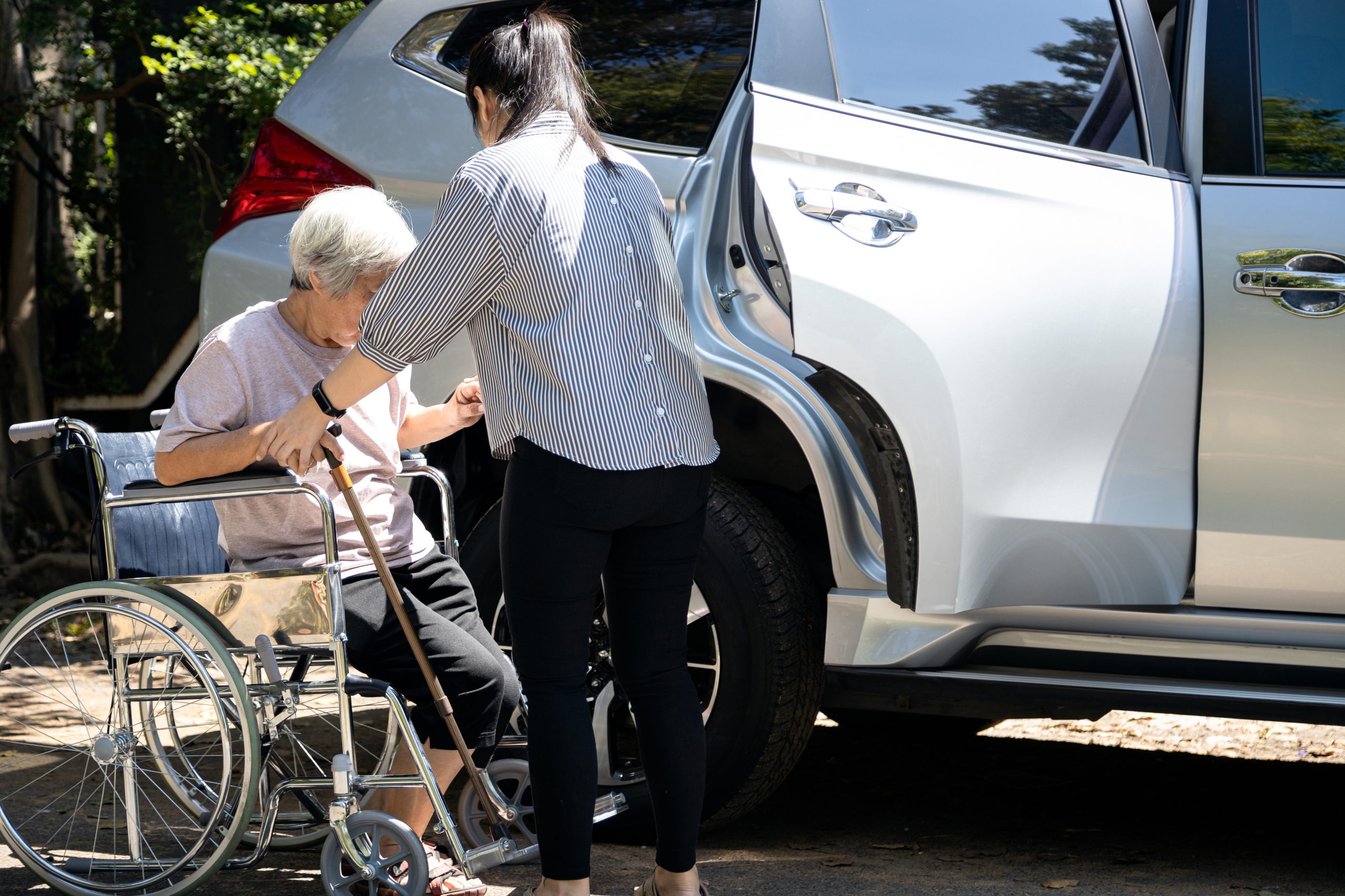 Asian female caregiver helping disabled elderly woman in wheelchair to get into the car,helpful daughter care and support senior mother to stand up from wheelchair in outdoor, caring for old people