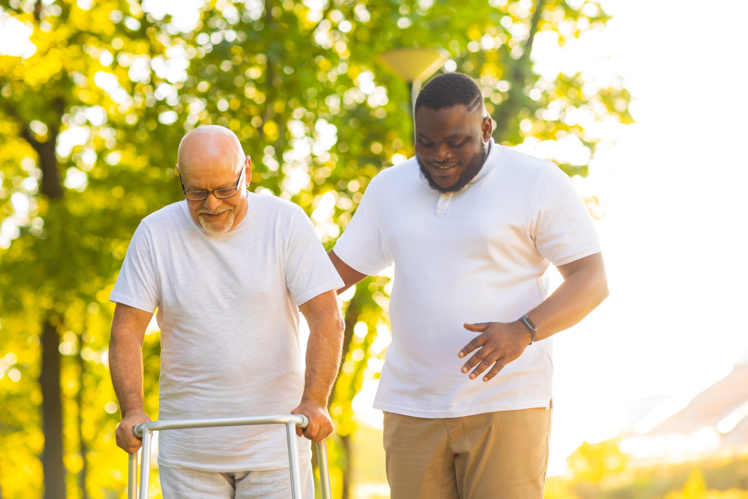 Caregiver is teaching old man to walk with walker. Professional nurse and patient walking outdoor at sunset. Assistance, rehabilitation and health care concept.