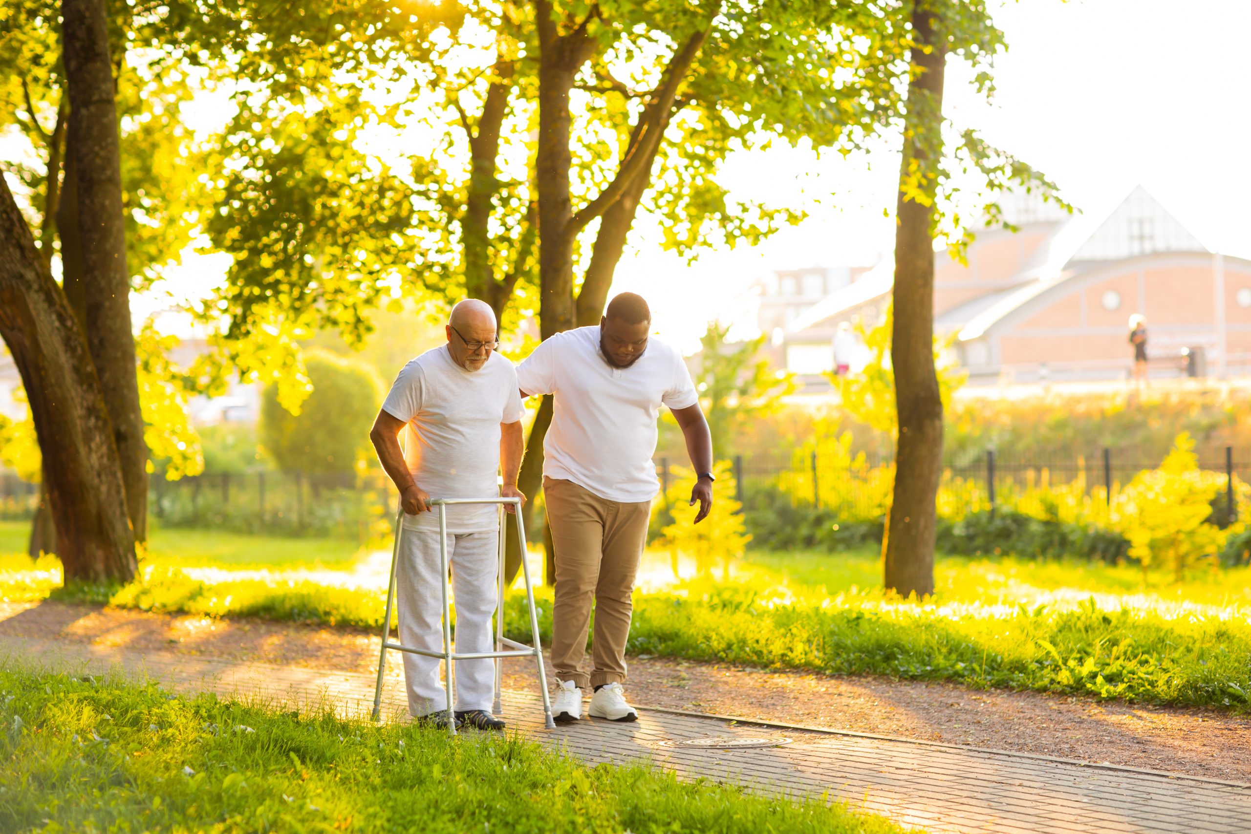 Caregiver is teaching old man to walk with walker. Professional nurse and patient walking outdoor at sunset. Assistance, rehabilitation and health care concept.